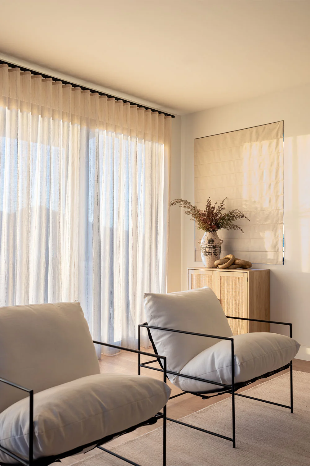 Bright contemporary living room with sheer Ripplefold drapery across a large window, two white cushioned black-frame chairs, and a neutral Roman shade.