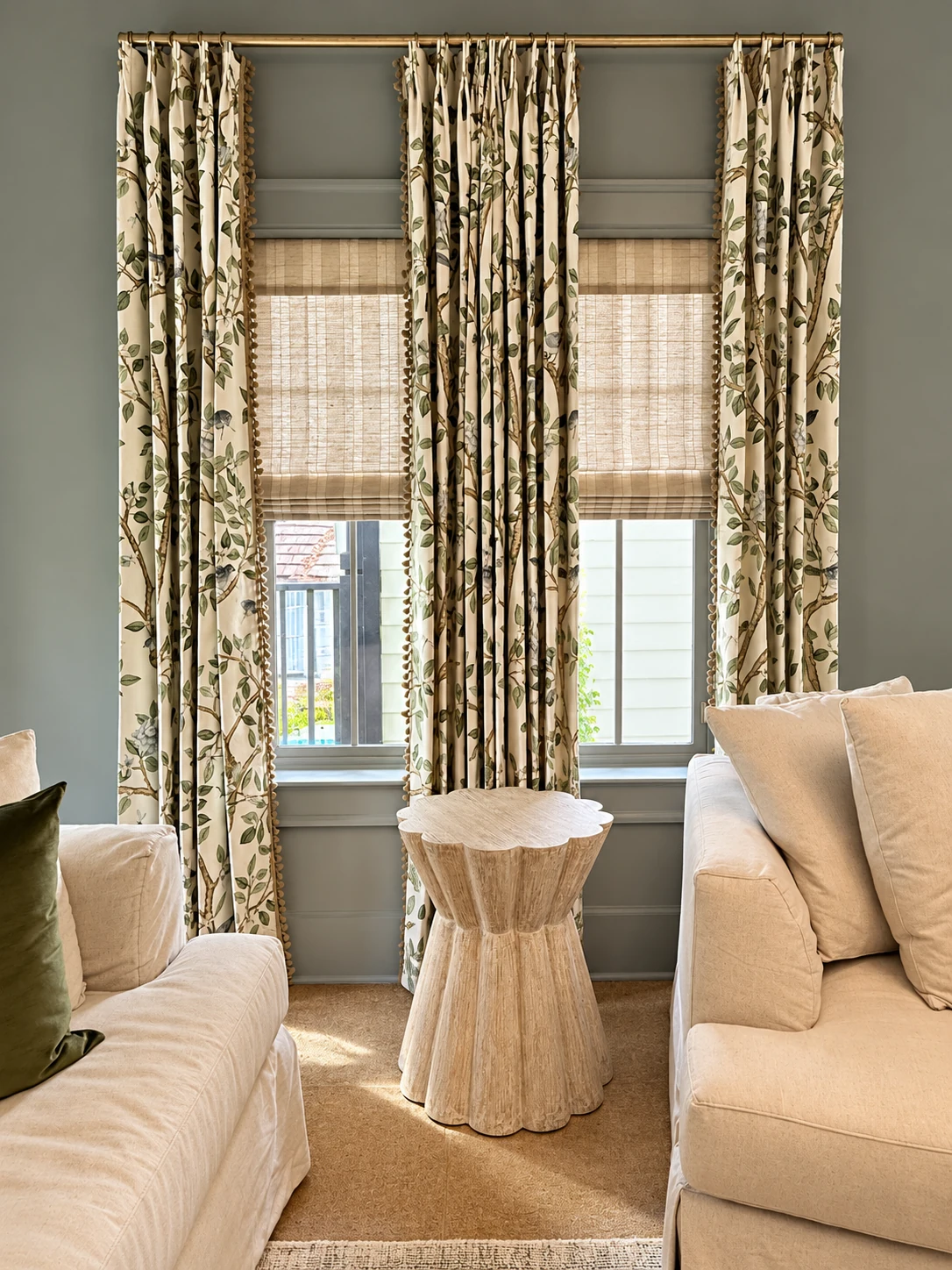 Botanical drapery layered with woven shades on brass hardware in a living room
