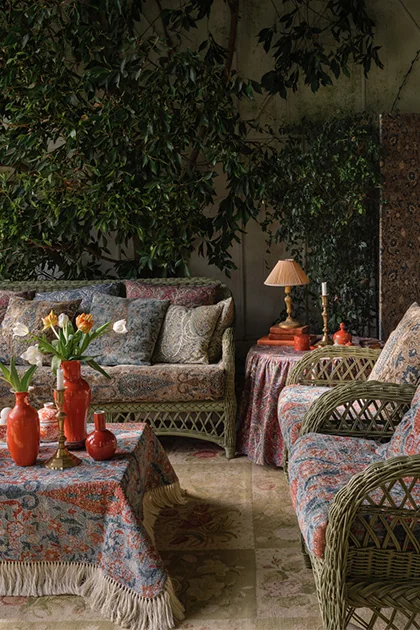 Travers textile-covered seating and tables in a richly styled leafy interior, showing patterned upholstery and fringed fabric surfaces with wicker chairs, a lamp, and botanical decor.