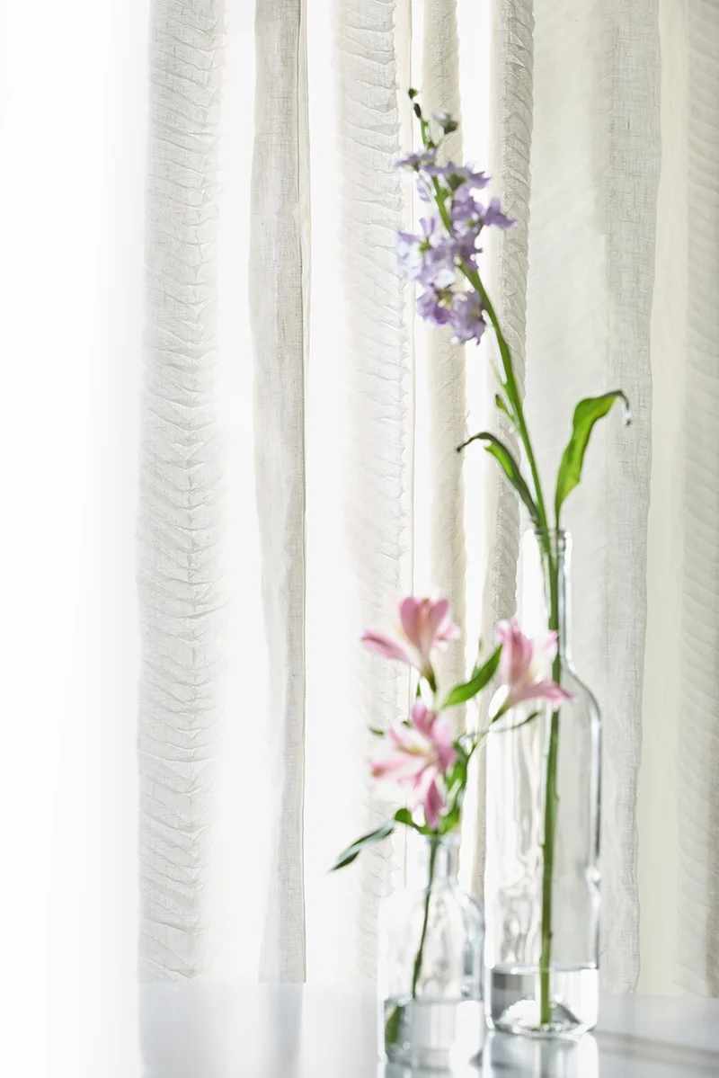Close-up styled photograph of Vervain Secret Garden sheer curtains with textured vertical fabric panels and flowers in glass vases.